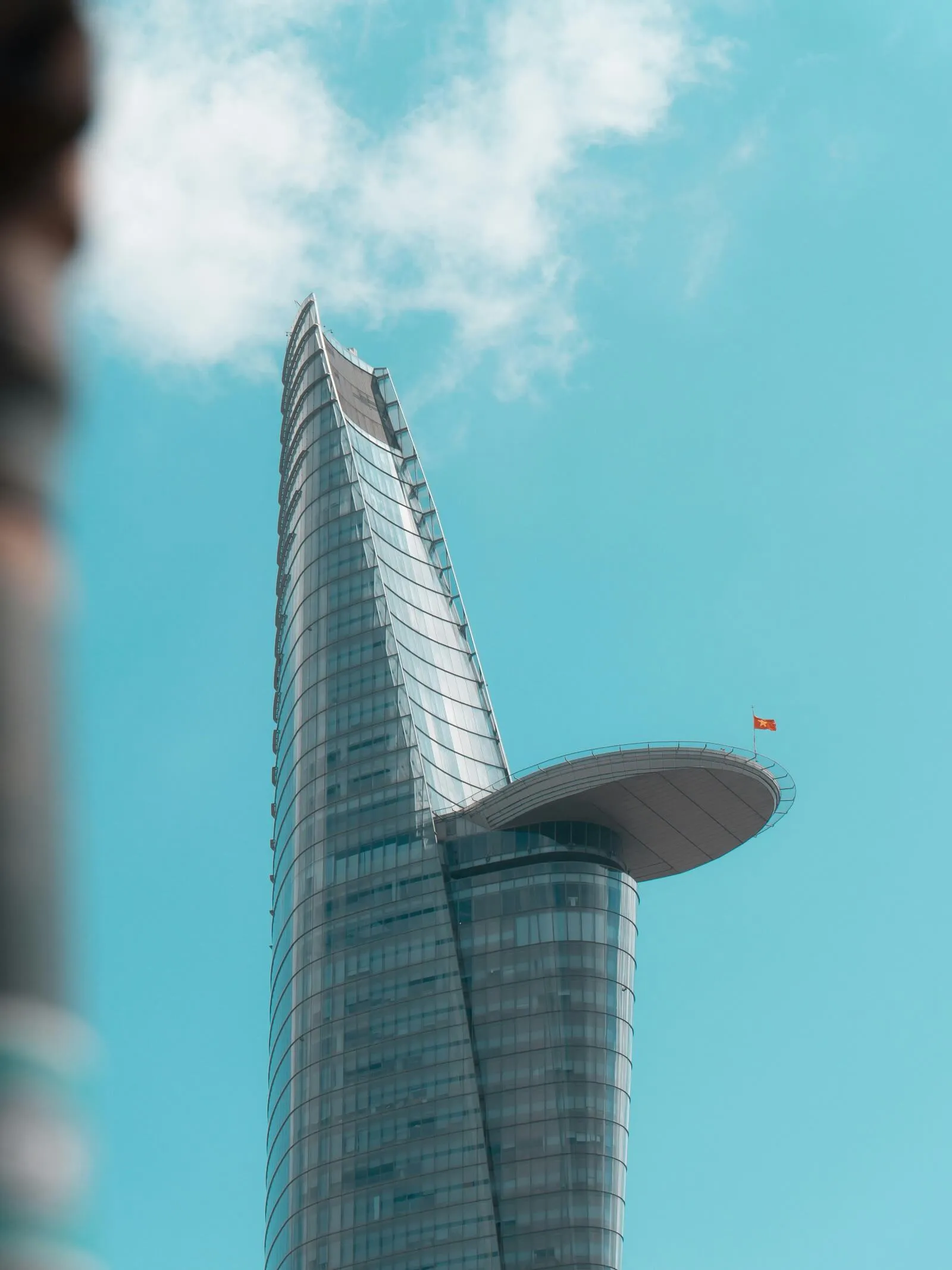 The top of Bitexco Financial Tower against a blue sky, with the helipad ledge and a Vietnamese flag jutting from its side